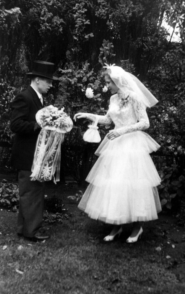 black and white image bride and groom standing in garden with dark backdrop of bushes. Groom handing bride bouquet.