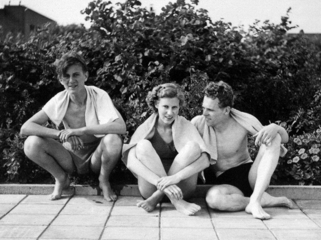 black and white image two men on either side of woman all wearing bathing suits and towels sitting on pavement in front of bushes