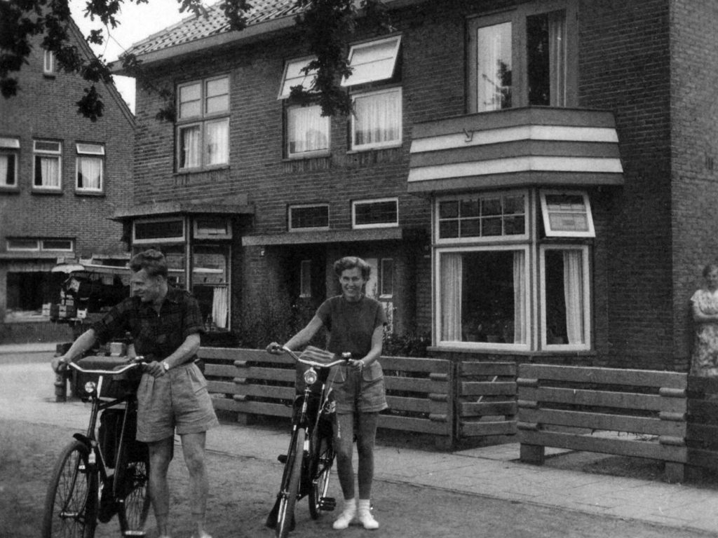 black and white image young couple wearing shorts standing with bicycles in street in front of two story brick townhouses