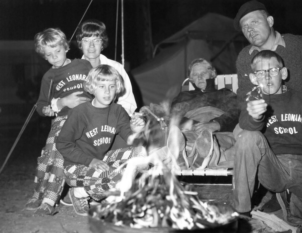 black and white image of mother father and three children wearing matching sweatshirts, older woman in background sitting in camping chair