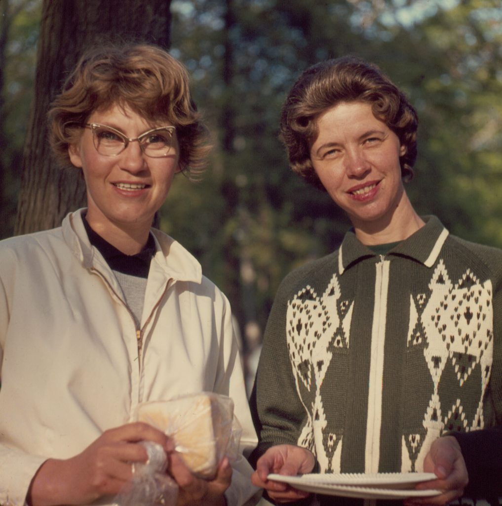 color image of woman with curled hair and 1960s glasses smiling and standing next to another woman with hair curled away from face